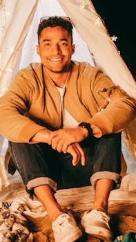 A cheerful man enjoying a night at the beach, sitting in a tent with warm string lights.