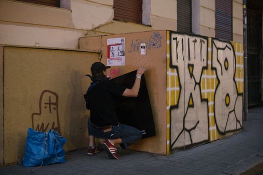 A street artist squats while adding graffiti to a wall on an urban street corner.