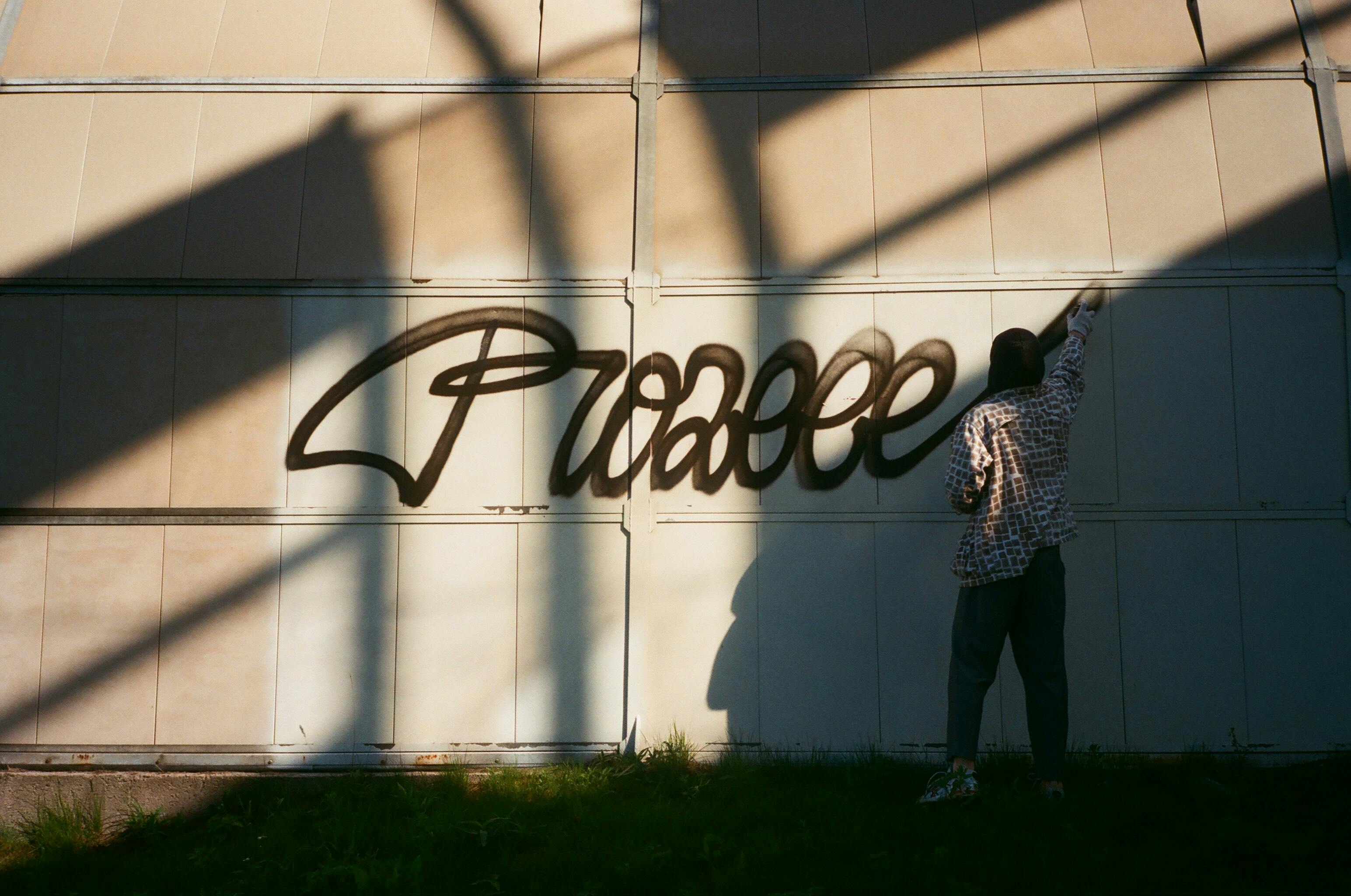 Street artist paints graffiti on wall in daylight, casting a shadow.