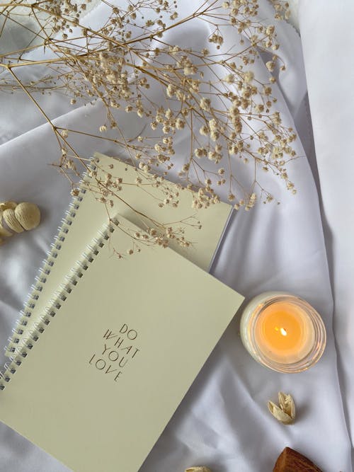 Free Top view composition of white spiral copybooks with inscription Do What You Want placed on white cloth with burning candle and tender flower Stock Photo