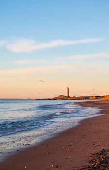 A picturesque view of a Danish beach at sunset with a distant lighthouse on the horizon.