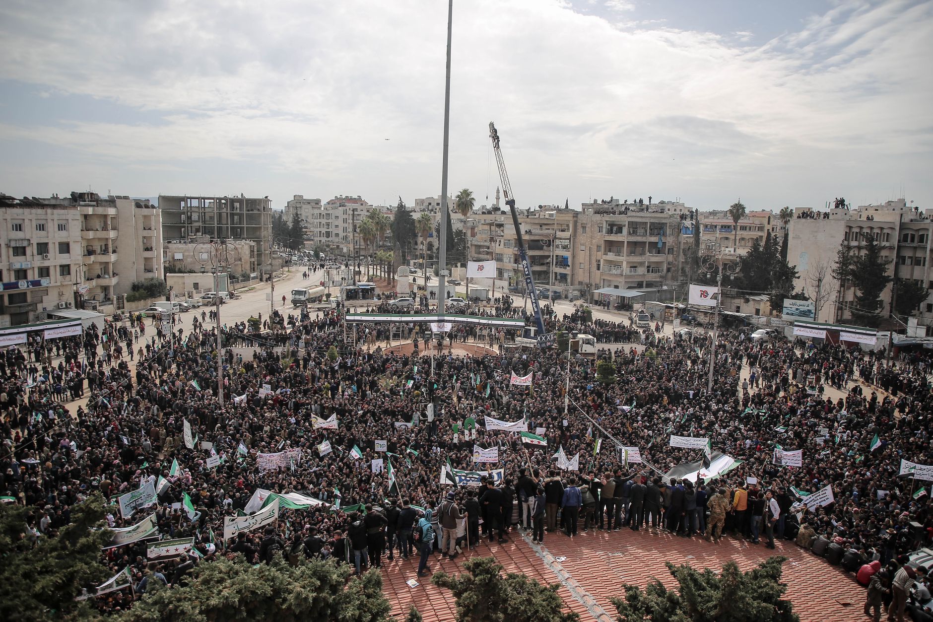 Aerial view of a large protest gathering in Idlib, Syria, with banners and signs.