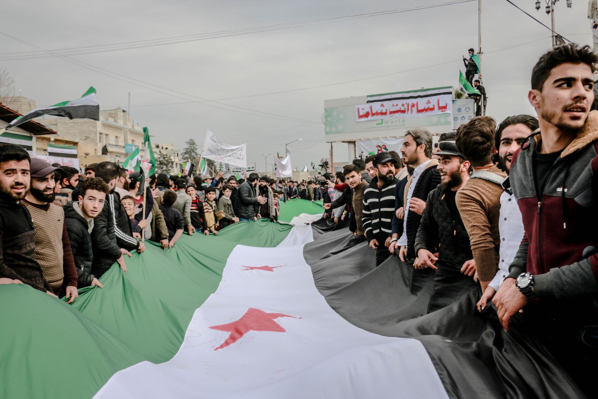 A mass protest in Idlib, Syria with demonstrators holding a large Syrian flag.