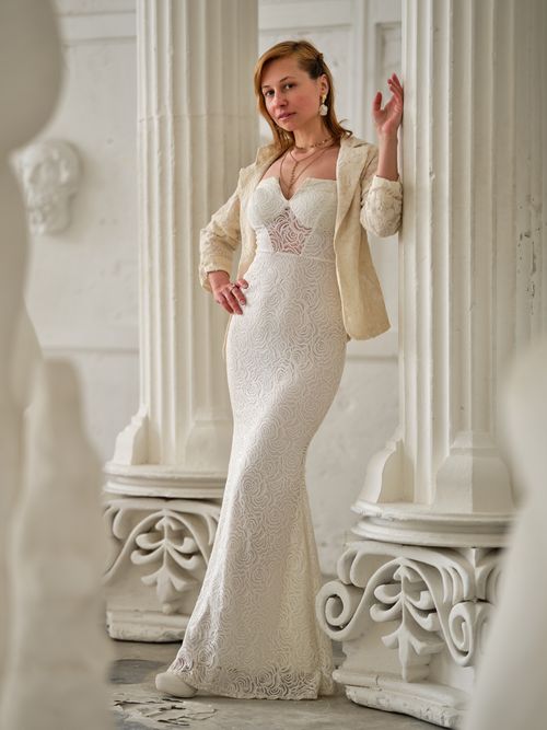 Free Full body of slender female in jacket and long white dress leaning on column in hall with sculptures Stock Photo