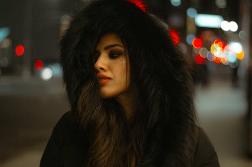 Charming female wearing black fur coat with hood looking away while standing on street with glowing lights on blurred background in evening time