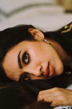 Face of attractive female with makeup and black hair looking away with serious glance while lying on bed on blurred background