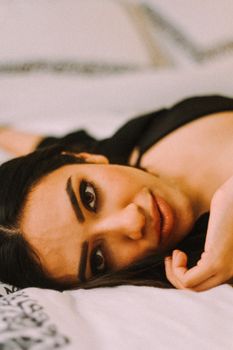 Positive female with black hair looking at camera while lying on soft bed in bedroom on blurred background at home