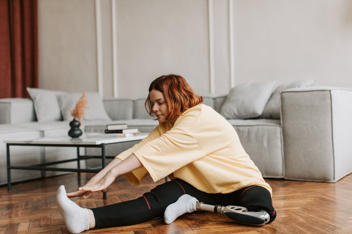 Free A woman with a prosthetic leg stretches on a wooden floor in her living room, embracing fitness. Stock Photo