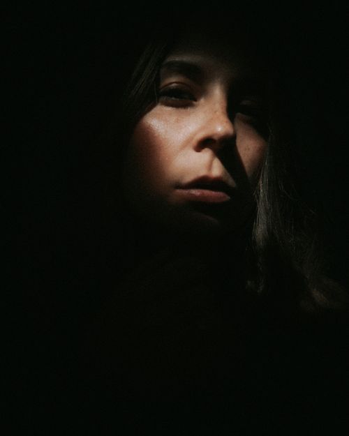 Free Face of unrecognizable mysterious female covered with shadow looking at camera while standing in dark room on black background with faint beam of light Stock Photo