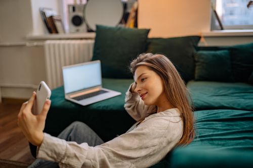Free Young woman relaxing on a couch while taking a selfie indoors with a laptop nearby. Stock Photo