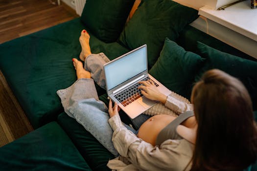 Pregnant woman sitting comfortably on a sofa using a laptop, working remotely.