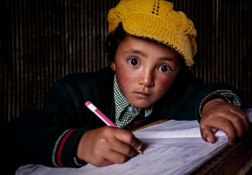 A young child intensely focused on writing indoors, wearing a yellow hat and green sweater.