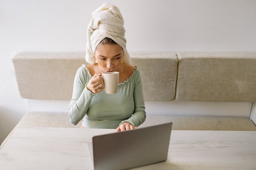 Relaxed woman with head towel sipping tea while using a laptop at home.