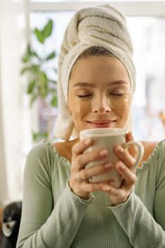 A woman with eye patches and towel relaxes while holding a warm cup of tea indoors.