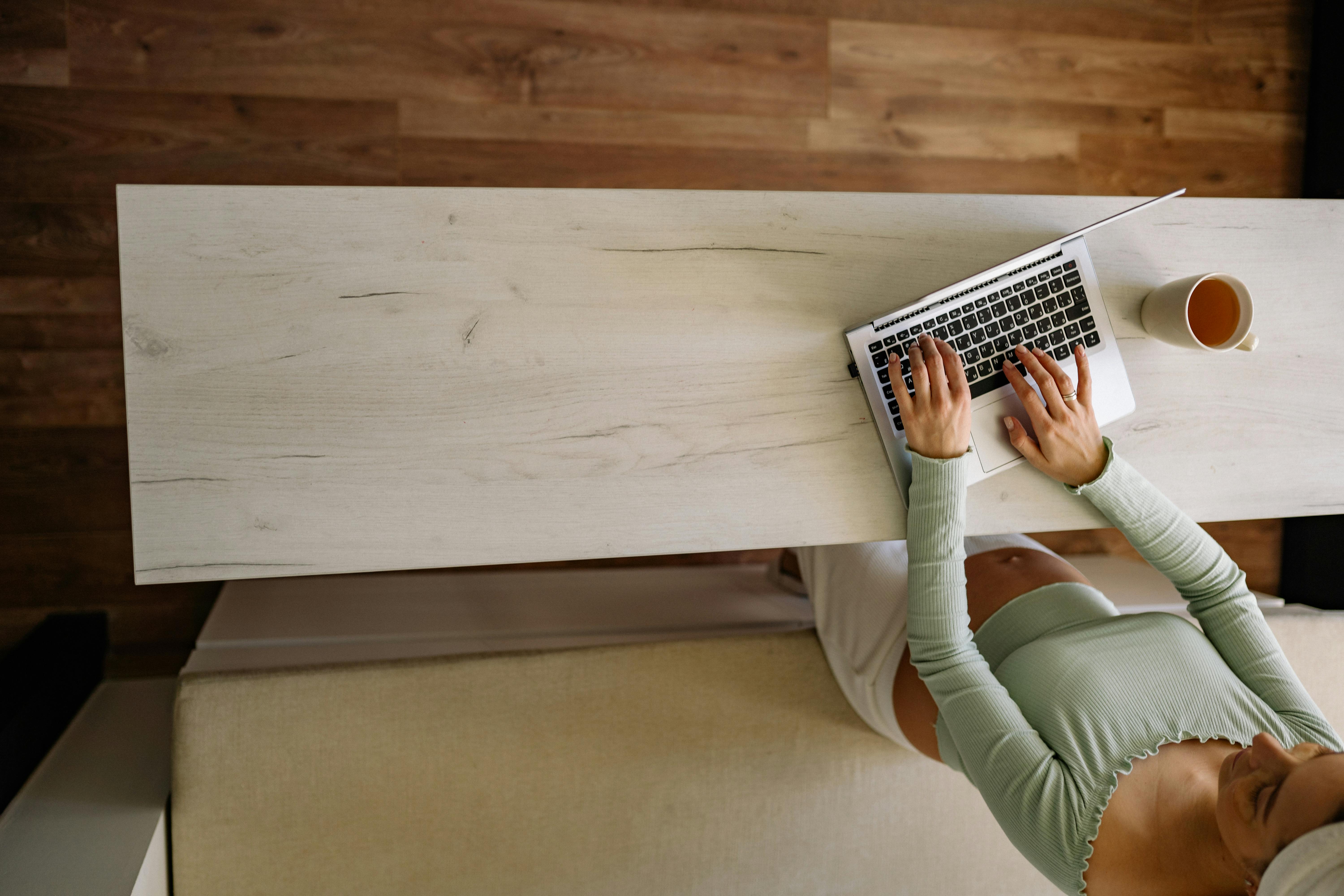 A pregnant woman working remotely from home, using a laptop with a coffee nearby.