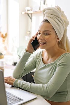 Smiling woman sitting indoors, talking on the phone with a head towel and under-eye patches.