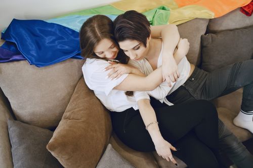 Free Two women embrace lovingly on a sofa with a rainbow flag, symbolizing pride and affection. Stock Photo