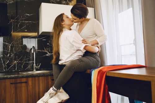 Free Affectionate same-sex couple embracing in a contemporary kitchen setting with an LGBT pride flag. Stock Photo