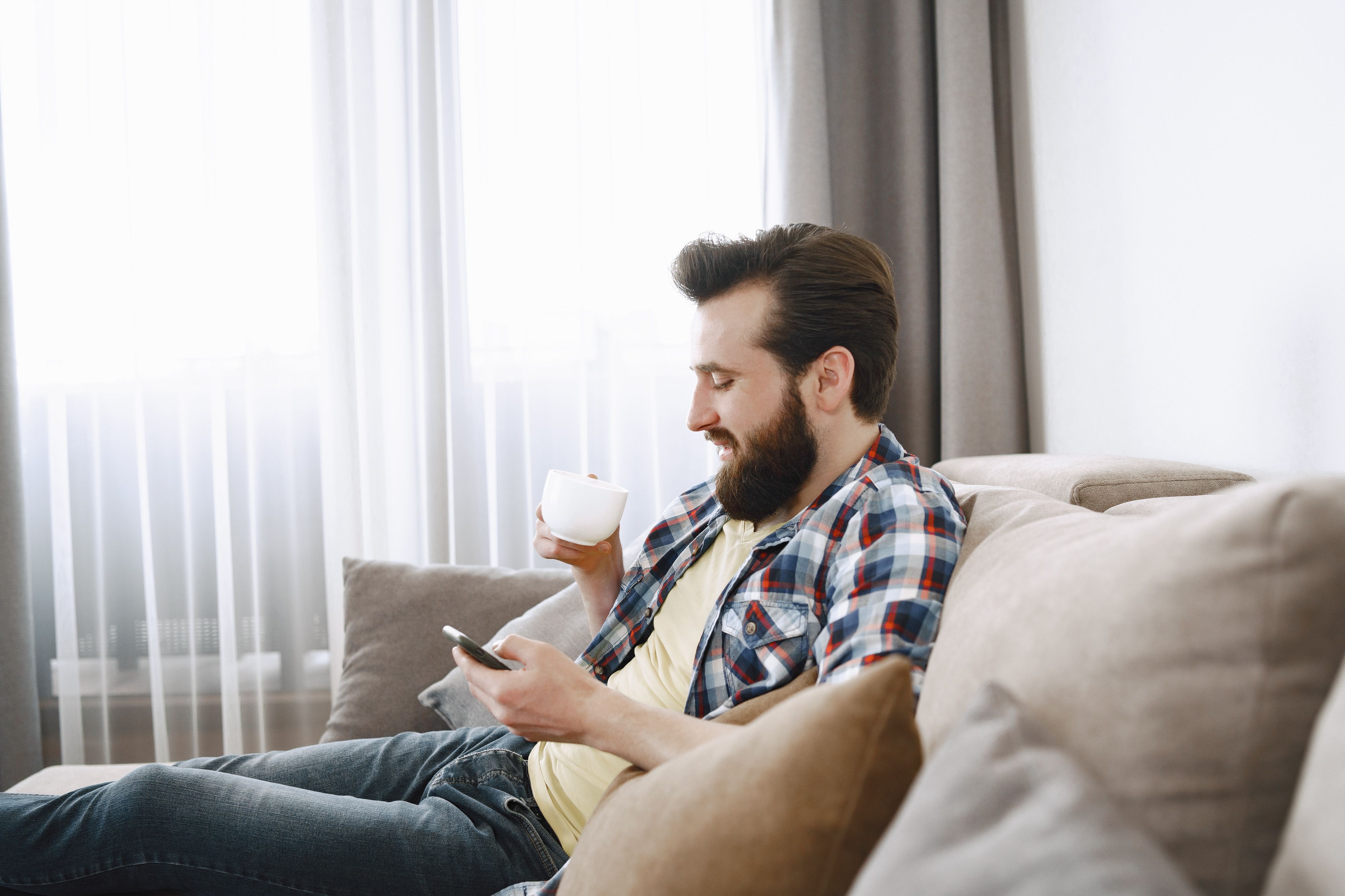 Casual bearded man enjoying coffee on a comfy sofa while browsing on his phone.