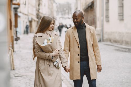 Free A multiracial couple walks hand in hand down a charming winter street, holding a bouquet. Stock Photo