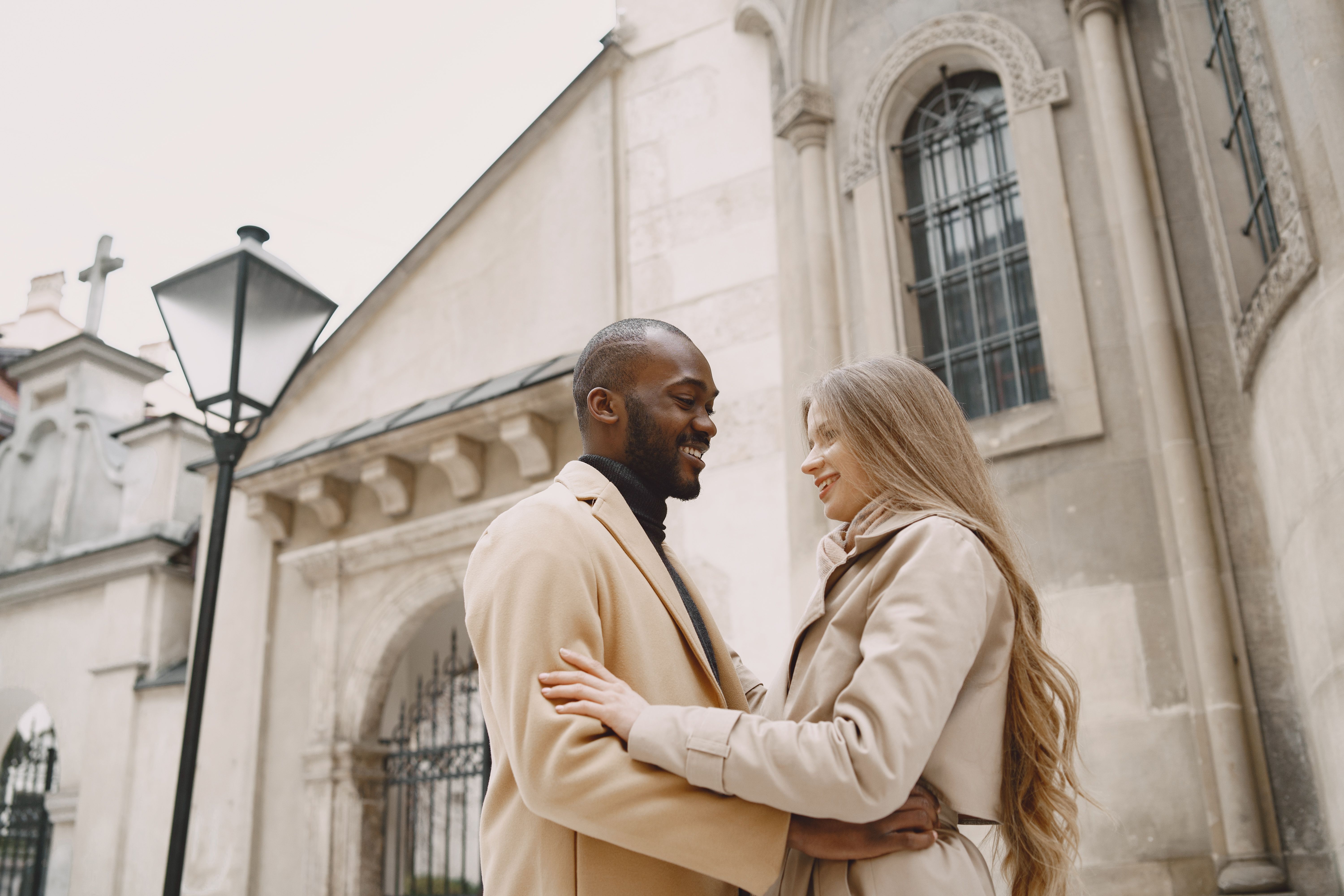 Diverse couple smiling and hugging near beautiful historic architecture, winter vibe.