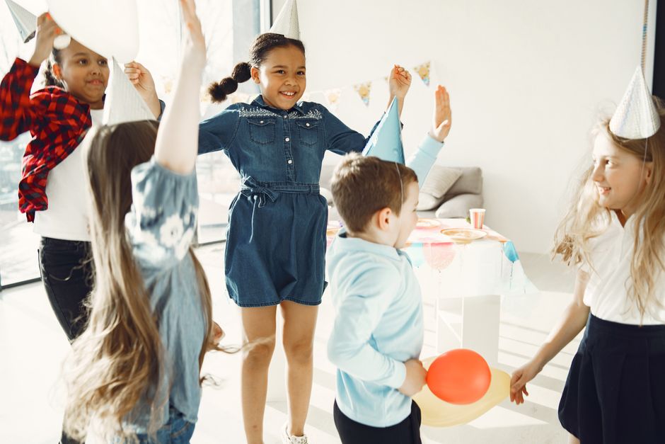 Children in party hats celebrating a birthday indoors with balloons and laughter.