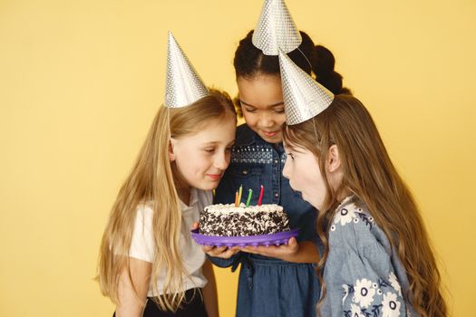 Three girls celebrate a birthday with hats and cake, against a yellow backdrop.