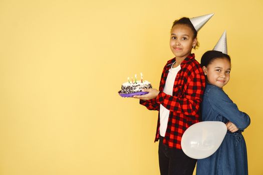 Two children celebrating a birthday with cake and balloons against a yellow background.