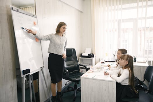 A female teacher guiding two girls during a lesson in a bright classroom with a whiteboard.