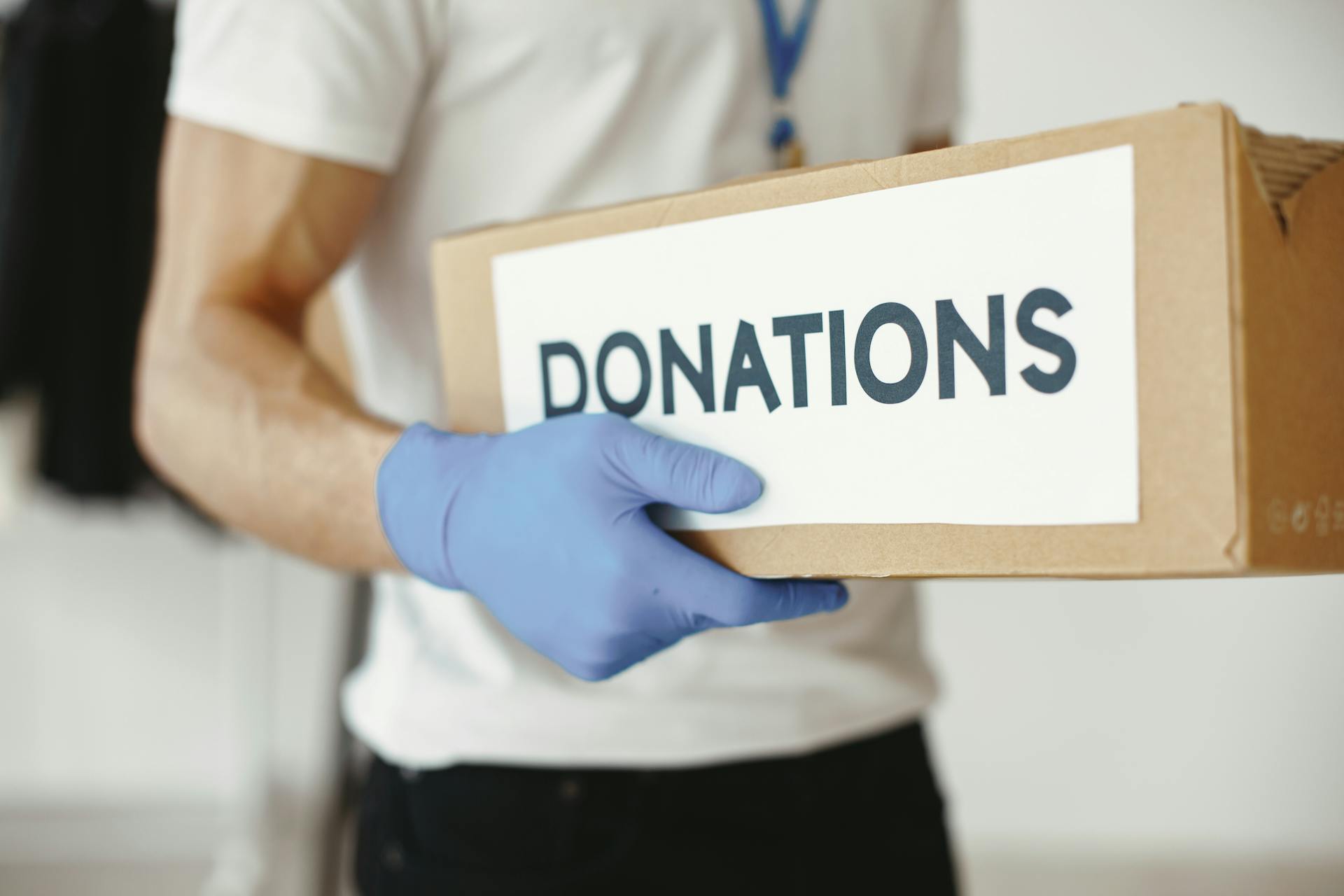 A volunteer wearing gloves holds a 'donations' box, symbolizing community support.