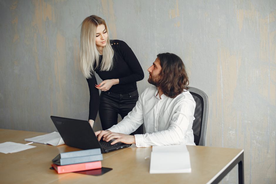 Two professionals in a modern office setting discussing work over a laptop with documents on the table.