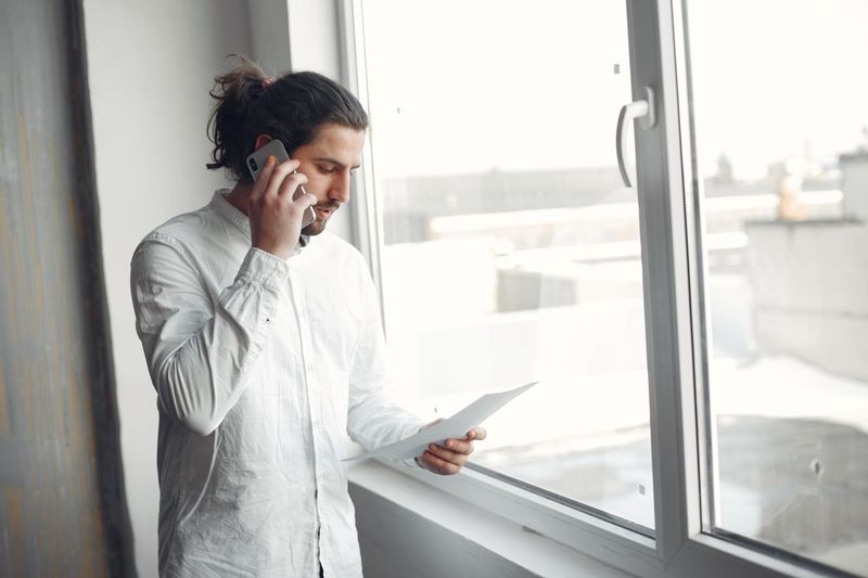 Young man in white shirt, on phone call holding a document, standing by a large window.