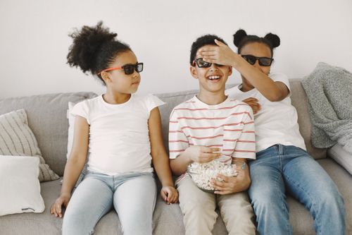 Free Three children having fun indoors with sunglasses and popcorn, enjoying leisure time on the sofa. Stock Photo