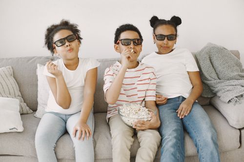 Free Three children watching a 3D movie on a sofa with popcorn, wearing 3D glasses indoors. Stock Photo