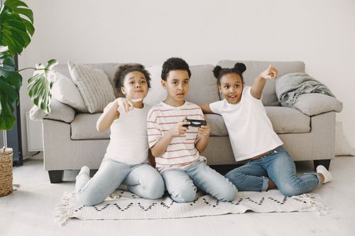 Free Children sitting on a rug, playing video games and pointing excitedly in a cozy living room. Stock Photo