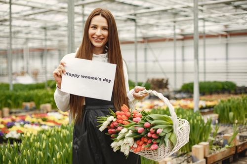 Free Woman with long hair smiling, holding flowers and a sign for women's day in a vibrant greenhouse. Stock Photo