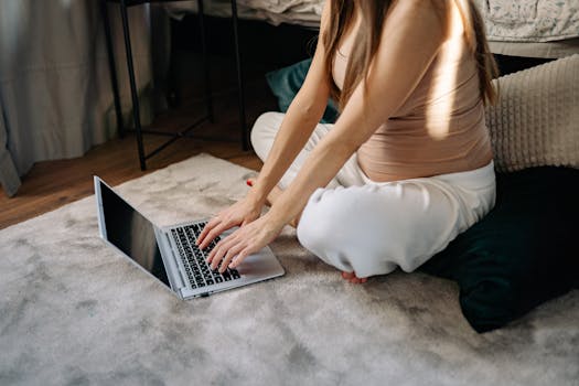 Pregnant woman working remotely on a laptop in a comfortable, sunlit bedroom area.