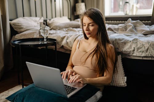 Free A Pregnant Woman Sitting on the Floor Leaning on a Bed while Busy Working on her Laptop Stock Photo