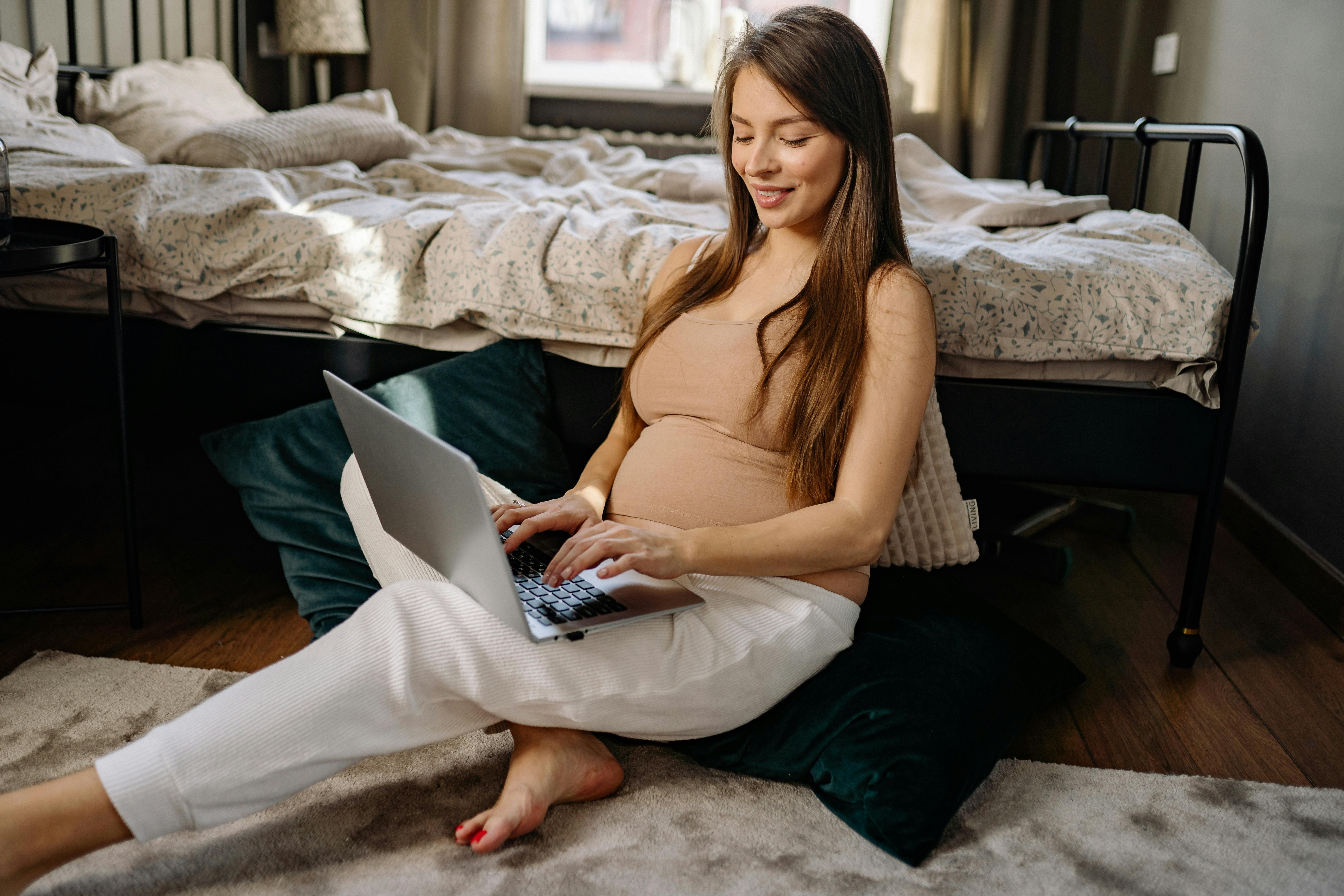 Pregnant woman using laptop, working remotely from home bedroom setting.