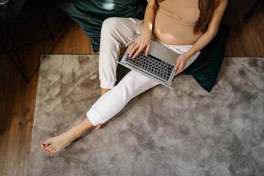 Pregnant woman sitting on carpet working remotely on laptop, enjoying a comfortable home environment.