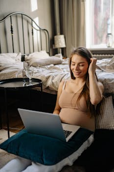 Pregnant woman smiling while working remotely on a laptop in a cozy bedroom setting.