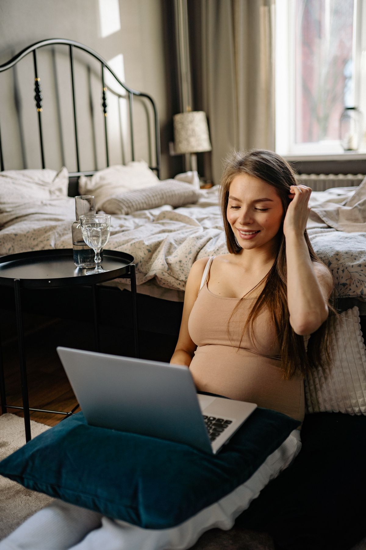 Pregnant woman smiling while using a laptop in a bright bedroom