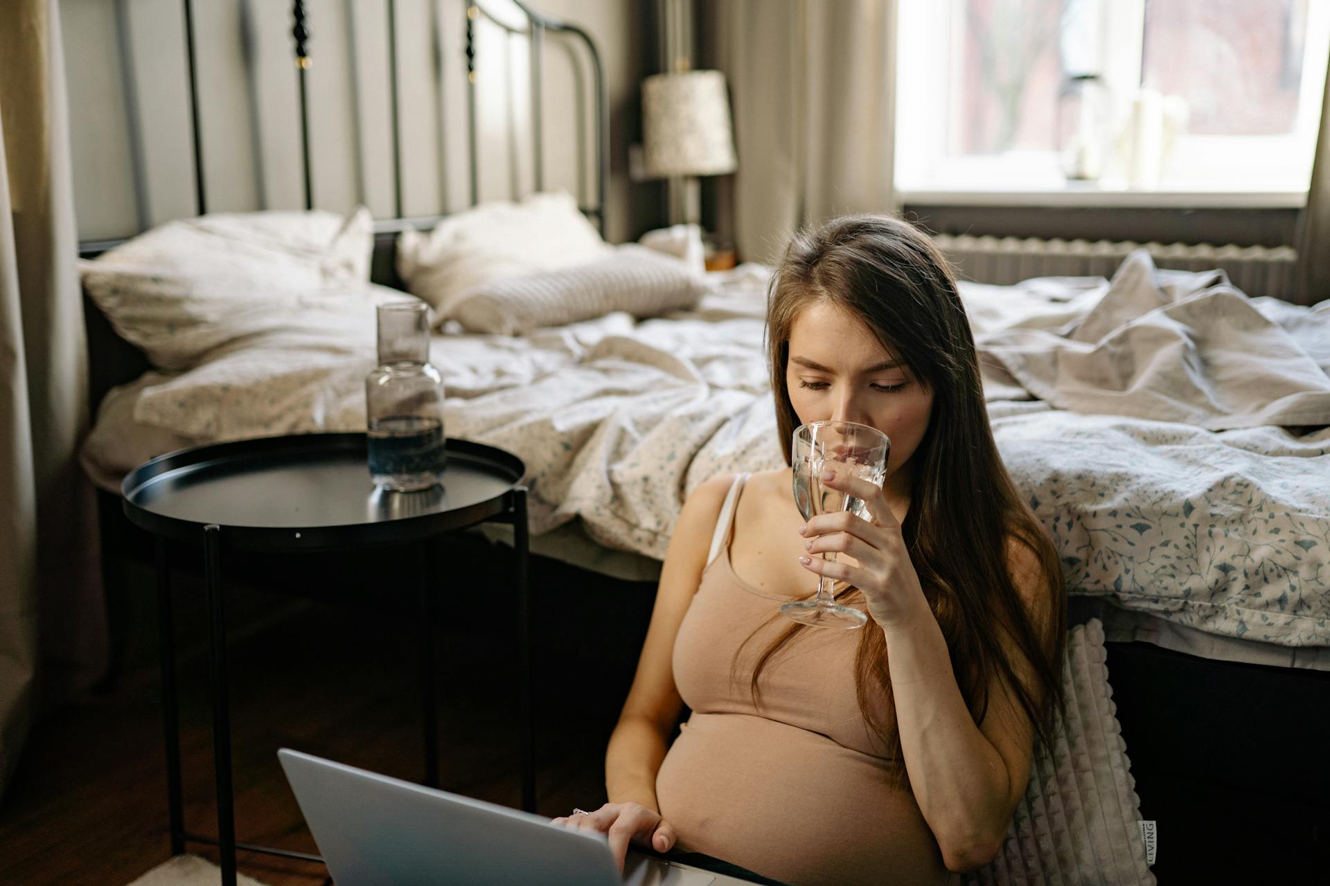 Pregnant woman sitting in a bedroom working on a laptop and drinking water.