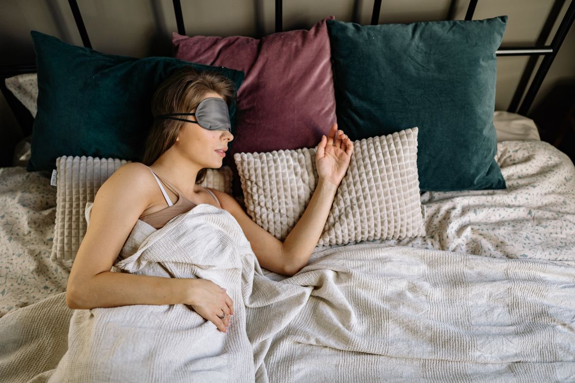 Woman asleep on her back in a gray eye mask, muted pastel pillows