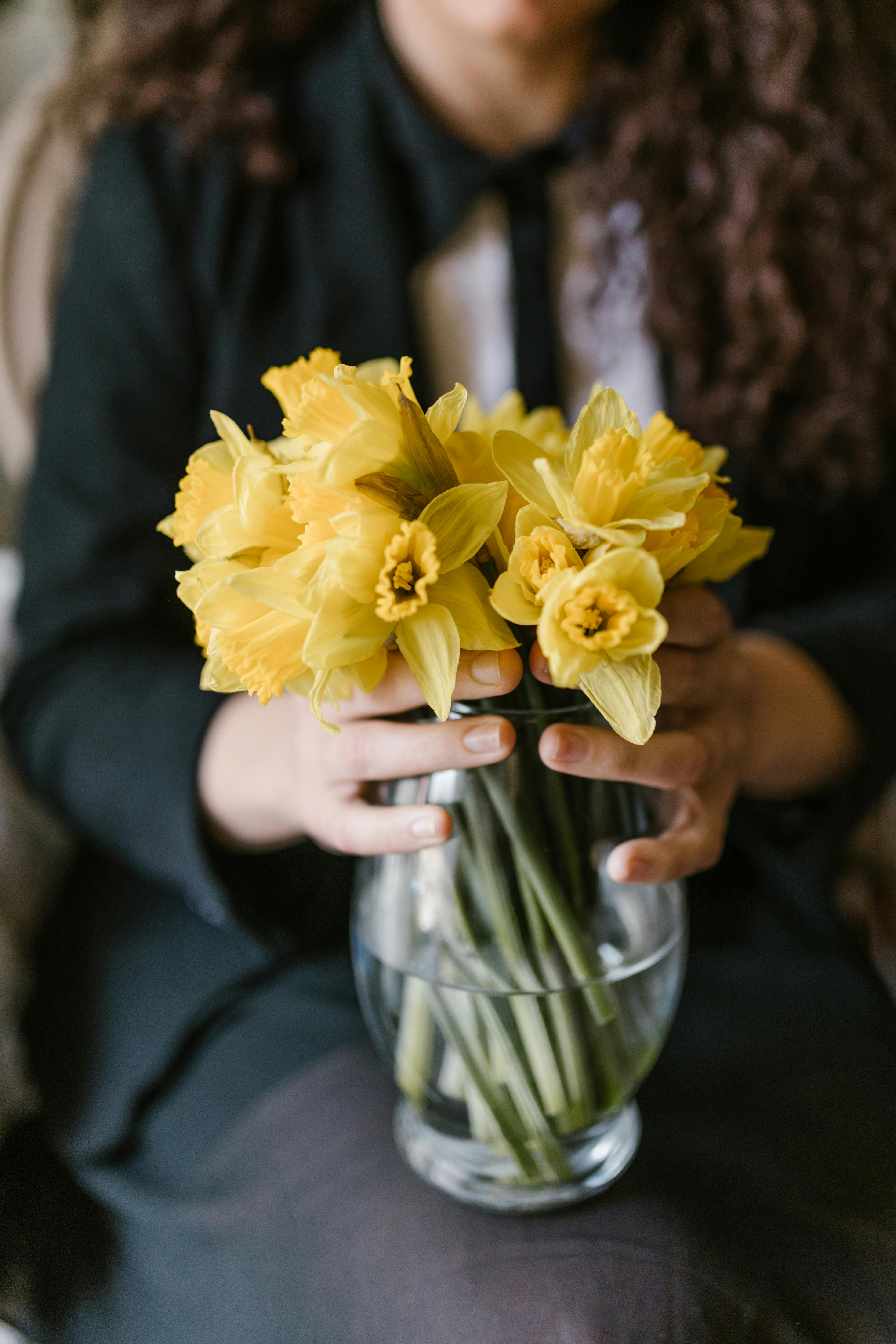 A woman holding a bouquet of yellow daffodils in a glass vase, indoors.
