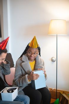 African American girl in party hat unwrapping gift during cozy indoor birthday celebration.