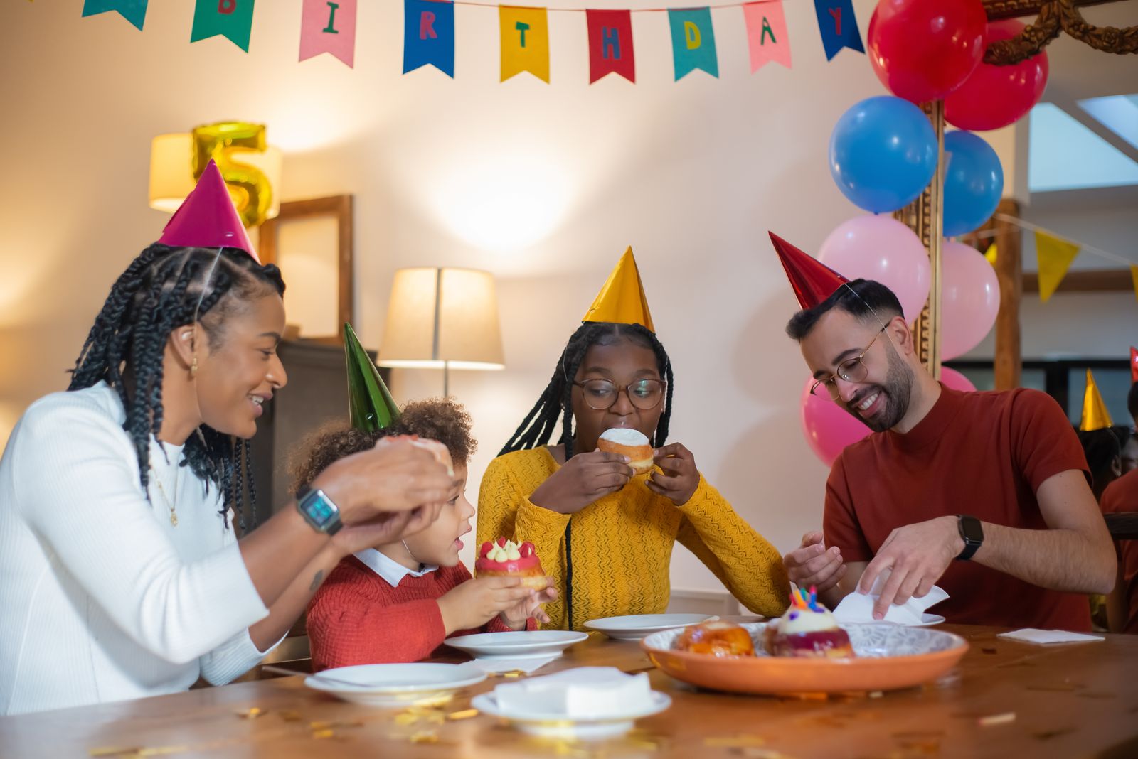 Children and adults wearing party hats sit at a birthday table and eat individual pastries.