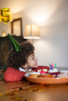 A joyful child wearing a party hat celebrates their fifth birthday indoors with a cake.
