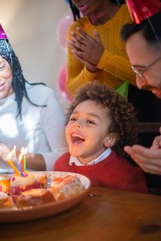 A happy child celebrates their birthday with family indoors, surrounded by cake and candles.