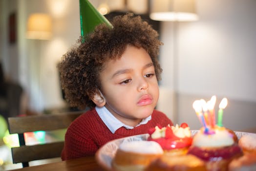 A thoughtful child in a party hat looks at candles on a birthday cake indoors.